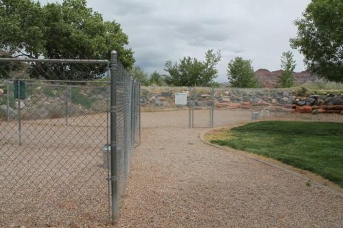 A fenced area with gravel and grass, featuring a gate and trees in the background under a cloudy sky.