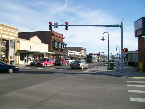 A busy street intersection with traffic lights, shops, and cars in a small town setting under a clear blue sky.