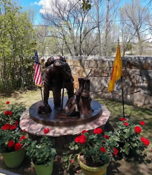 A bronze statue of a soldier kneeling, surrounded by flowers and flags, in a peaceful outdoor setting.