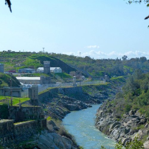 A river flows beside a fenced facility, with green hills and blue sky in the background.