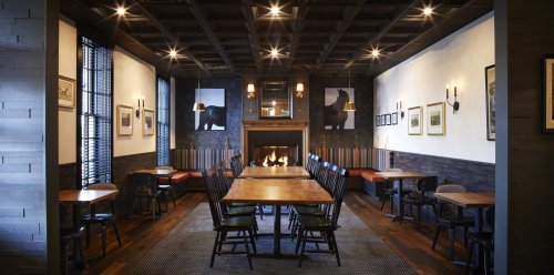 Cozy restaurant interior with wooden tables, chairs, and a fireplace, featuring artwork on the walls and warm lighting.