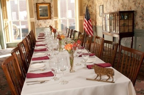 A long dining table set with glassware and flowers, in a vintage room with floral wallpaper and an American flag.
