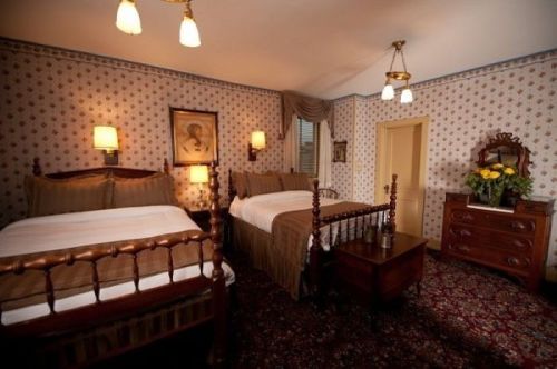 Cozy bedroom with two wooden beds, floral wallpaper, vintage decor, and a dresser with flowers. Warm lighting enhances ambiance.