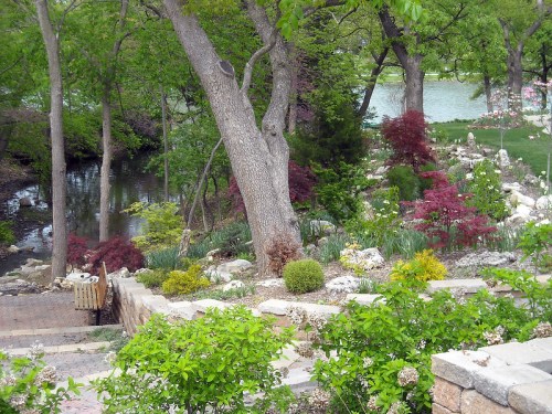A serene garden scene with lush greenery, colorful shrubs, and a tranquil pond in the background.