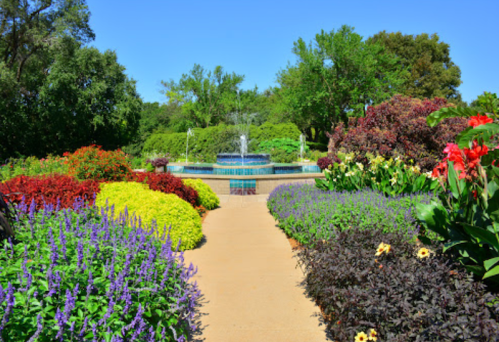 A vibrant garden path lined with colorful flowers leads to a fountain surrounded by lush greenery under a clear blue sky.