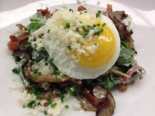 A fresh salad topped with a poached egg, crispy bacon, and grated cheese on a white plate.