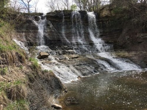 A serene waterfall cascading over rocky ledges into a calm pool, surrounded by greenery and trees.