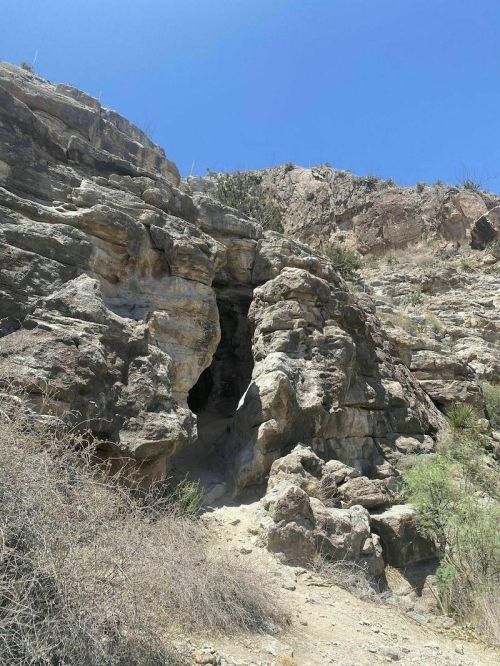 A rocky cave entrance surrounded by dry vegetation and steep hills under a clear blue sky.