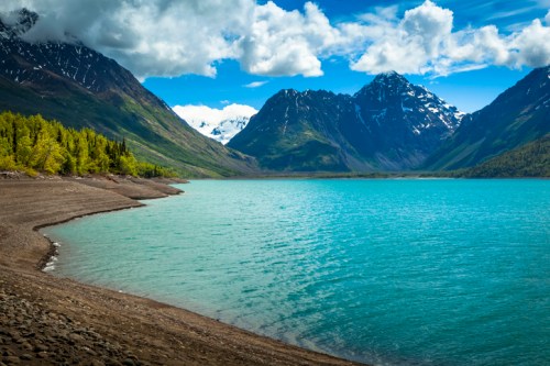 A serene lake surrounded by mountains, with clear blue water and lush green trees under a partly cloudy sky.