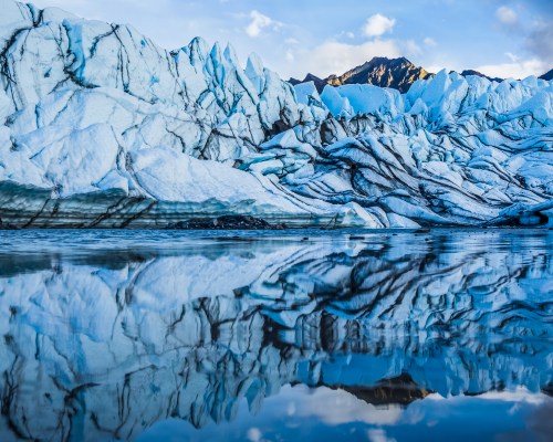 A stunning glacier with jagged ice formations reflecting in calm water, surrounded by mountains under a clear blue sky.