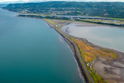 Aerial view of a coastal landscape with a narrow road separating the ocean from a river and lush green fields.