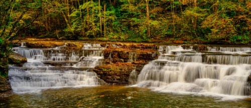 A serene waterfall cascades over rocky ledges, surrounded by lush green trees and autumn foliage.