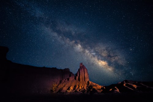 A stunning night sky filled with stars and the Milky Way above a rugged rock formation illuminated by soft light.
