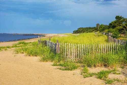A sandy beach with tall grass, a wooden fence, and a calm waterway under a cloudy sky.