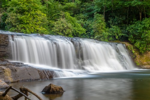 A serene waterfall cascades over rocks into a calm pool, surrounded by lush green trees and foliage.