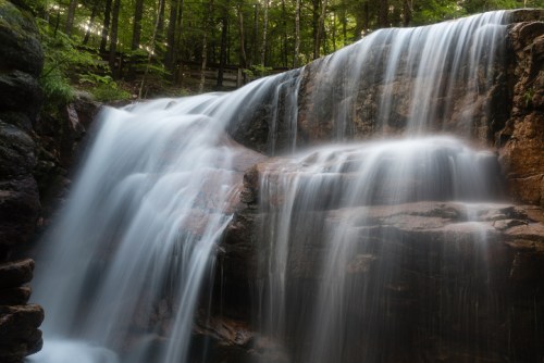 A serene waterfall cascades over rocky ledges, surrounded by lush green trees in a tranquil forest setting.