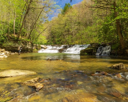 A serene waterfall cascades into a clear stream, surrounded by lush green trees and rocky banks under a blue sky.