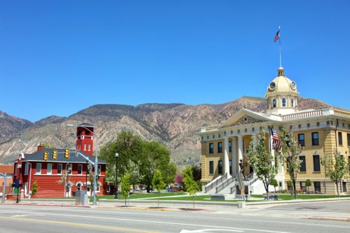 A historic courthouse with a dome stands next to a red building, set against a backdrop of mountains and a clear blue sky.