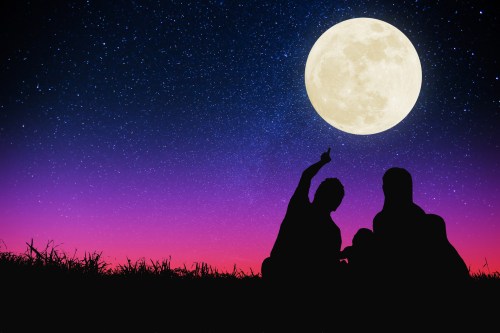 Silhouettes of two people sitting on grass under a starry sky with a large full moon above.