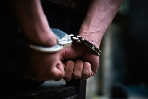 Close-up of hands in handcuffs, gripping a wooden surface, conveying a sense of restraint and confinement.