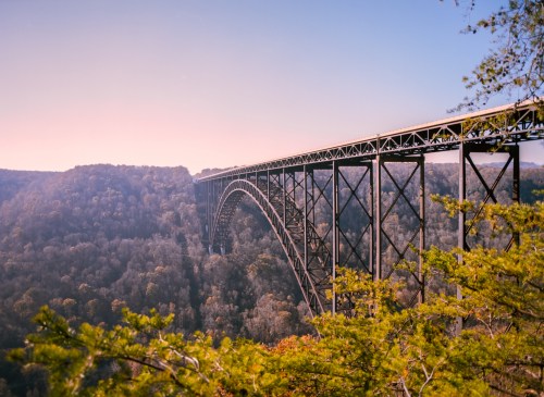 A large metal bridge arches over a valley filled with trees, under a pastel sky at sunset.