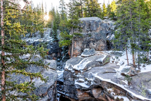 A rocky landscape with snow, surrounded by evergreen trees, and sunlight peeking through the forest.