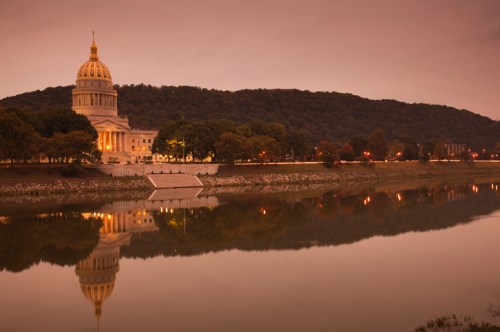 A serene view of a capitol building reflecting in a river at dusk, surrounded by trees and hills.