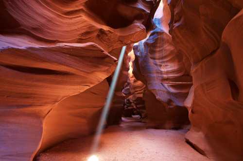A narrow canyon with smooth, wavy walls and a beam of light illuminating the sandy floor.