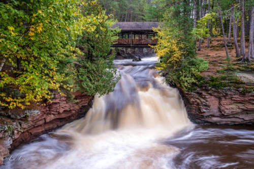 A serene waterfall cascades over rocky cliffs, surrounded by lush trees and a wooden bridge in the background.