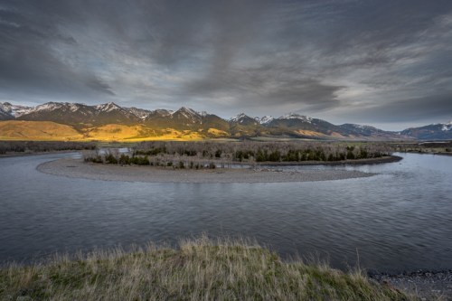 A winding river flows through a valley, surrounded by mountains under a cloudy sky at dusk.