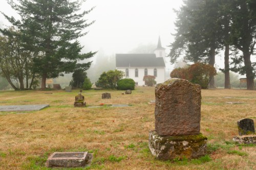 A foggy cemetery with gravestones in the foreground and a white church in the background, surrounded by trees.