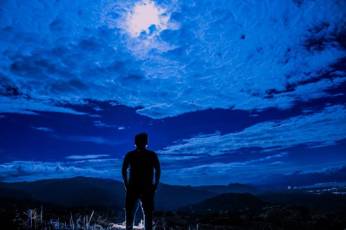 Silhouette of a person standing on a hill under a bright moon and dramatic clouds at night.