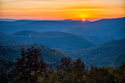 A vibrant sunset over rolling mountains, with layers of trees and hills silhouetted against the colorful sky.