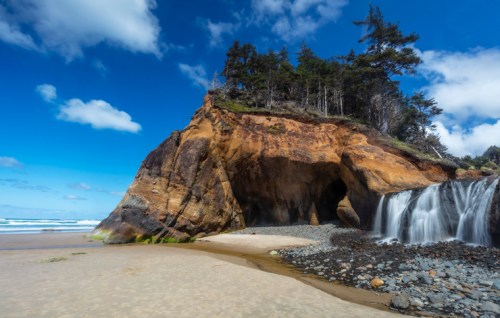 A rocky coastal cliff with trees, a waterfall, and a sandy beach under a blue sky with scattered clouds.
