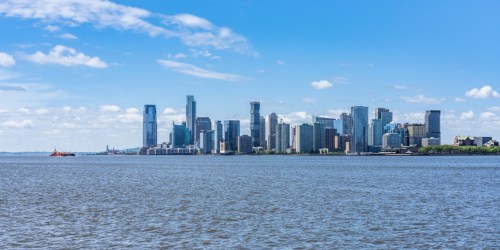 A panoramic view of a city skyline with tall buildings against a blue sky, reflecting on a calm river.