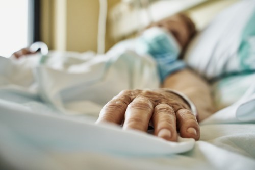A close-up of a patient's hand resting on a hospital bed, with a blurred background of a hospital room.