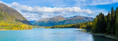 A serene lake surrounded by mountains and lush trees under a blue sky with fluffy clouds.