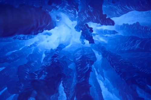 Abstract view of blue ice formations resembling stalactites, illuminated from above, creating a mystical underwater effect.
