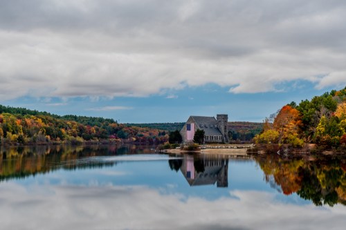 A serene lake reflects a stone building with a tower, surrounded by vibrant autumn foliage and cloudy skies.