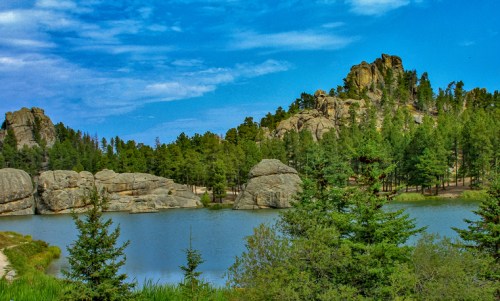A serene lake surrounded by lush trees and rocky formations under a blue sky with scattered clouds.