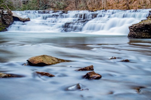 A serene waterfall cascades over rocks into a calm river, surrounded by trees and autumn foliage.