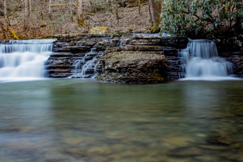 A serene waterfall cascades over rocky ledges into a calm pool, surrounded by trees and natural greenery.