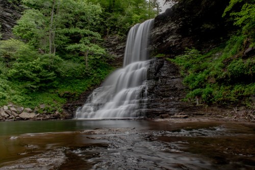A serene waterfall cascades over rocky cliffs, surrounded by lush green trees and a calm pool below.