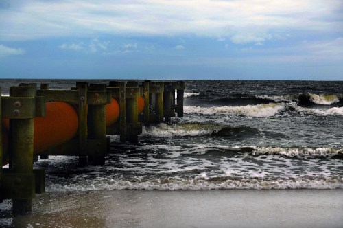 A weathered pier extends into a dark ocean, with gentle waves lapping at the sandy shore under a cloudy sky.