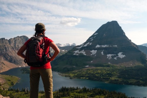 A hiker stands on a mountain overlook, gazing at a towering peak and a serene lake below, surrounded by lush greenery.