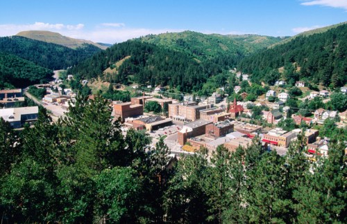 A scenic view of a small town nestled in green hills, surrounded by trees and mountains under a clear blue sky.