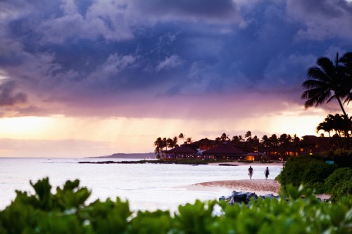 A serene beach at sunset with two people walking along the shore, palm trees, and dramatic clouds overhead.