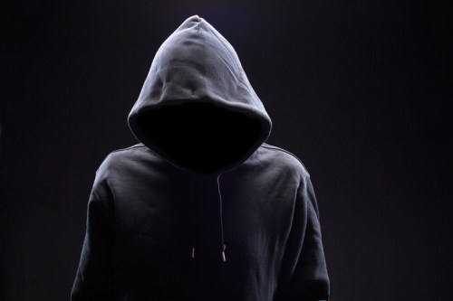 A figure in a dark hoodie stands against a black background, with the face obscured by shadows.