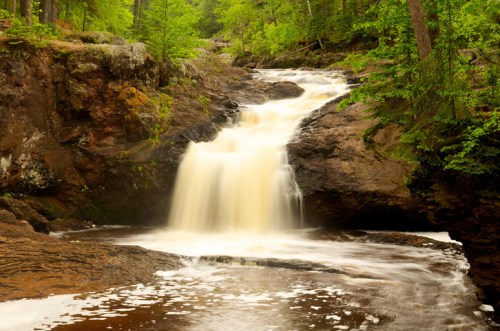 A serene waterfall cascades over rocky terrain, surrounded by lush green trees and a tranquil pool below.