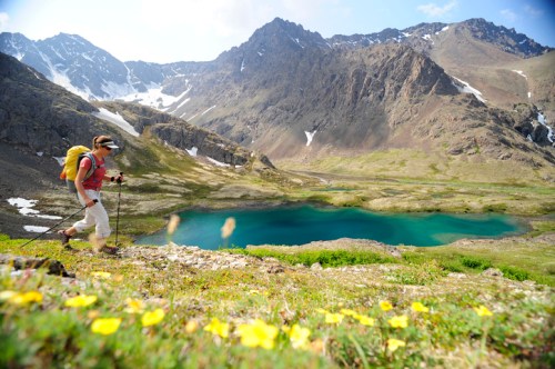 A hiker walks along a mountain trail with a lake and wildflowers in the foreground, surrounded by rugged peaks.